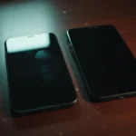 Dois celulares com a tela apagada lado a lado sobre mesa de madeira escura